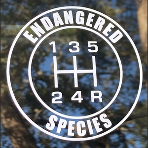 May include: A black and white sticker with the text "ENDANGERED SPECIES" in a circle around a gear shift pattern with the numbers 1, 2, 3, 4, 5, and R.