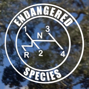 May include: White sticker with the words "ENDANGERED SPECIES" in a circular design. The design includes a gear shift pattern with numbers and letters. The background is a blurred view of trees and sky.