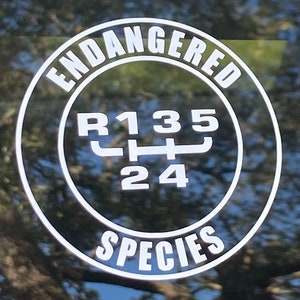 May include: A white circle sticker with the words "Endangered Species" around the outside. Inside the circle is a black and white gear shift pattern with the numbers "R135" and "24".