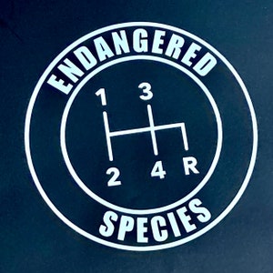 May include: A black and white sticker with the text "Endangered Species" in a circle around a white gear shift pattern with the numbers 1, 2, 3, 4, and the letter R.