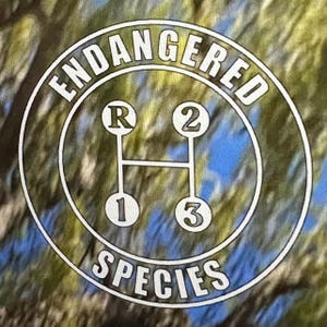 May include: A t-shirt with a circular graphic featuring the words "ENDANGERED SPECIES" around the edge. Inside, a white gear shift pattern shows R, 1, 2, and 3. The background has a camouflage pattern of green, blue, and brown.