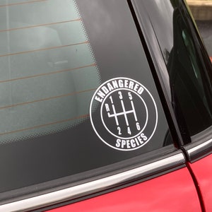 May include: A white vinyl sticker on a red car window. The sticker is a circle with a black outline and the words "Endangered Species" written in white. Inside the circle is a black and white illustration of a car's gear shift with the numbers 1, 2, 3, 4, 5, and 6.