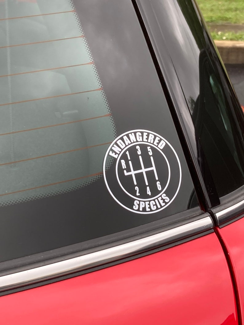 May include: A white vinyl sticker on a red car window. The sticker is a circle with a black outline and the words "Endangered Species" written in white. Inside the circle is a black and white illustration of a car's gear shift with the numbers 1, 2, 3, 4, 5, and 6.