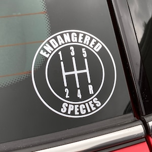 May include: A black and white sticker with the text "Endangered Species" in a circle around a gear shift pattern. The numbers 1, 2, 3, 4, 5, and R are on the gear shift pattern.