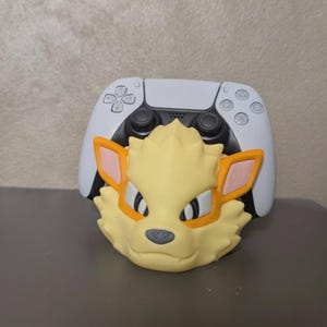 Puede incluir: Un soporte para mando con temática de Arcanine amarillo y naranja con un mando blanco de PlayStation 5 en el fondo. La figura de Arcanine tiene una cara detallada y está hecha de un material sólido.