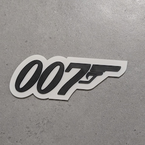 Peut inclure: Un autocollant noir et blanc représentant le logo "007" et la silhouette d'une arme à feu. Le design a un effet en relief en trois dimensions, avec les chiffres et le pistolet en noir sur fond blanc.