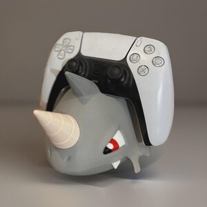 Può includere: Una statuina di rinoceronte grigio con un controller PlayStation 5 bianco e nero sopra. Il rinoceronte ha un corno bianco e dettagli rossi sul viso. Il controller presenta i simboli PlayStation.