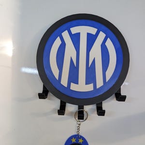 Op de afbeelding: Een rond, blauw en wit logo met een zwarte rand, met de letters "I", "M" en "T". Onder het logo bevinden zich vier zwarte haken. Een sleutelhanger met hetzelfde logo en twee gouden sterren hangt aan de haken.