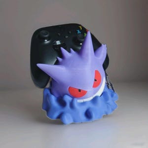 Puede incluir: Un soporte para mando de Gengar morado con ojos rojos y una sonrisa blanca, sosteniendo un mando de videojuegos negro. El soporte tiene una base azul en forma de nube. Un divertido accesorio para jugadores.