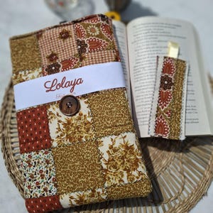 Puede incluir: Una funda de libro de tela de retazos con cierre de botón y un marcapáginas a juego. La funda presenta una variedad de patrones florales y geométricos marrones, beiges y rojos. La funda tiene la palabra "Lolaya" impresa en una banda blanca.
