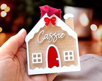 Peut inclure: Une décoration de Noël en forme de maison en pain d'épices en bois avec un toit blanc et une porte rouge. La décoration est décorée de glaçage blanc et porte le nom "Cassie" sur le devant.