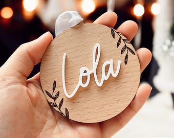 Peut inclure: Une décoration ronde en bois avec le nom "Lola" écrit en lettres cursives blanches. La décoration est ornée de motifs floraux noirs.