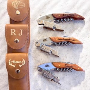 Könnte beinhalten: Personalisierte Weinöffner mit Holzgriffen und Edelstahlkomponenten. Die Öffner verfügen über eingravierte Namen und ein Traubenmuster. Eine braune Ledertragetasche ist ebenfalls enthalten.