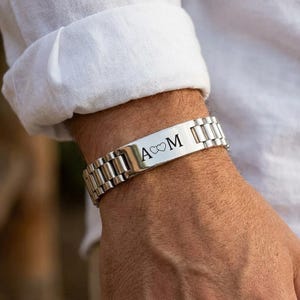 Puede incluir: Una pulsera de eslabones de metal plateado con una placa rectangular central grabada con la letra A, un símbolo de corazón infinito y la letra M. La pulsera tiene un acabado pulido y un diseño de banda estilo reloj con eslabones metálicos.