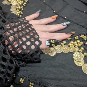 May include: A black fishnet sleeve with a scalloped edge worn over a black fabric adorned with gold-toned coins. The fingernails feature various designs, including one nail with a black base, white drip accents, and the gold letters GHD.