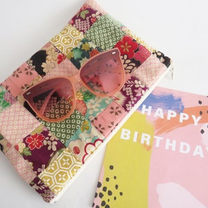 May include: A pink and white patchwork fabric clutch with a zipper closure and pink sunglasses on top. A pink and yellow birthday card with the text "Happy Birthday" is in the background.