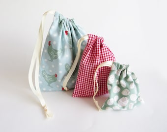 May include: Three drawstring bags made of fabric. The largest bag is light blue with white polka dots and a green chicken print. The middle bag is red and white gingham. The smallest bag is green with white polka dots.