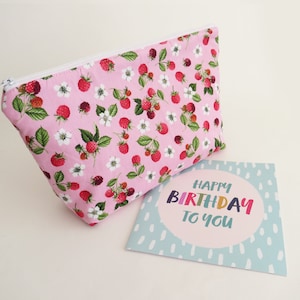 May include: A pink zippered pouch with a white zipper and a floral pattern of white flowers and red raspberries. A blue card with white polka dots and the text "Happy Birthday To You" in colorful letters.