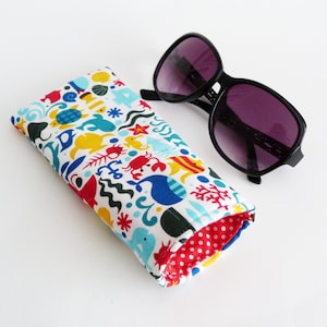 May include: A colorful fabric case with a sea life pattern, featuring fish, seahorses, and coral. The case is rolled up and has a red and white polka dot lining. A pair of black sunglasses with purple lenses is lying next to the case.