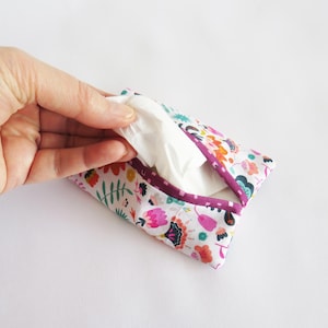 May include: A floral patterned tissue holder with a purple trim, held open to reveal white tissues. The fabric features a white background with colorful flowers and leaves. The holder is open, and tissues are being pulled out.