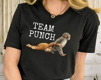 Op de afbeelding: Zwart T-shirt met de tekst "TEAM PUNCH" in het wit. Het shirt heeft een afbeelding van twee apen, een grijze en een bruine, waarbij de grijze aap een tak vasthoudt. De persoon draagt een blauwe spijkerbroek.