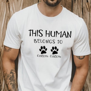 May include: White t-shirt with the text "This Human Belongs To" and two paw prints with the word "Custom" under each paw print.