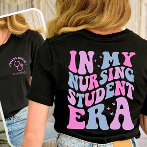 May include: Black t-shirt with the text "IN MY NURSING STUDENT ERA" in pink and blue retro font. The front of the shirt has the text "FUTURE NURSE" with a stethoscope graphic. A stylish tee for nursing students.