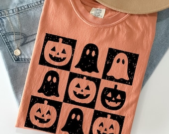 May include: A peach-colored t-shirt featuring a grid pattern of black jack-o'-lanterns and ghosts. The shirt is laid flat with a pair of light blue jeans and a tan hat. A gold necklace is visible.