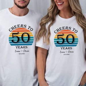 May include: Two white t-shirts with a retro sunset graphic and the text "Cheers to 50 Years". The shirts have the names "Emma" and "Dean" and the date "12.02.1950" printed on them.