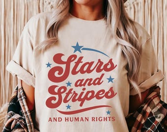 Könnte beinhalten: Beiges T-Shirt mit dem Text "Stars and Stripes AND HUMAN RIGHTS" in Rot und Blau. Das Shirt hat hochgekrempelte Ärmel und wird mit einem karierten Hemd kombiniert. Das Gesamtthema ist patriotisch und politisch.