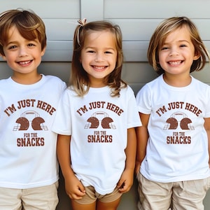 May include: Three children wearing white t-shirts with brown text that reads "I'm just here for the snacks" and a graphic of two football helmets. The children are wearing khaki shorts.