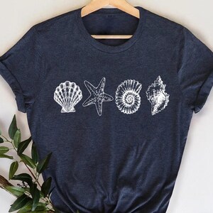 May include: A navy blue t-shirt featuring a white seashell graphic. The graphic includes a scallop shell, starfish, nautilus shell, and conch shell. The shirt is displayed on a wooden hanger.
