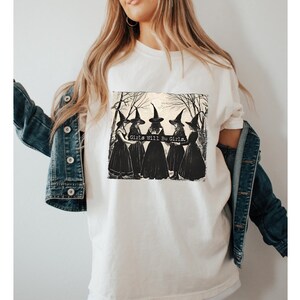 K&ouml;nnte beinhalten: Wei&szlig;es T-Shirt mit einem schwarz-wei&szlig;en Grafikdruck von vier Hexen in einem Wald. Der Text "Girls Will Be Girls" ist auf dem Grafikdruck abgebildet.