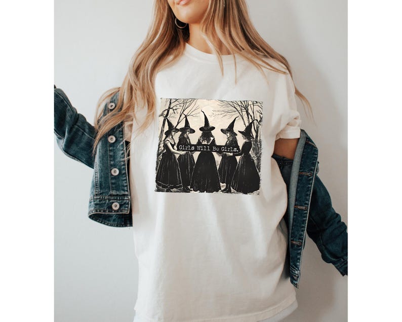 K&ouml;nnte beinhalten: Wei&szlig;es T-Shirt mit einem schwarz-wei&szlig;en Grafikdruck von vier Hexen in einem Wald. Der Text "Girls Will Be Girls" ist auf dem Grafikdruck abgebildet.
