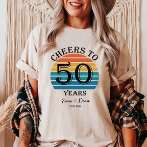 May include: A white t-shirt with a retro-style graphic that says "Cheers to 50 Years" with a rainbow-coloured sunset behind the number 50. The text "Emma &hearts; Dean 12.02.1950" is below the graphic.