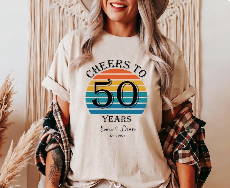 May include: A white t-shirt with a retro-style graphic that says "Cheers to 50 Years" with a rainbow-coloured sunset behind the number 50. The text "Emma &hearts; Dean 12.02.1950" is below the graphic.