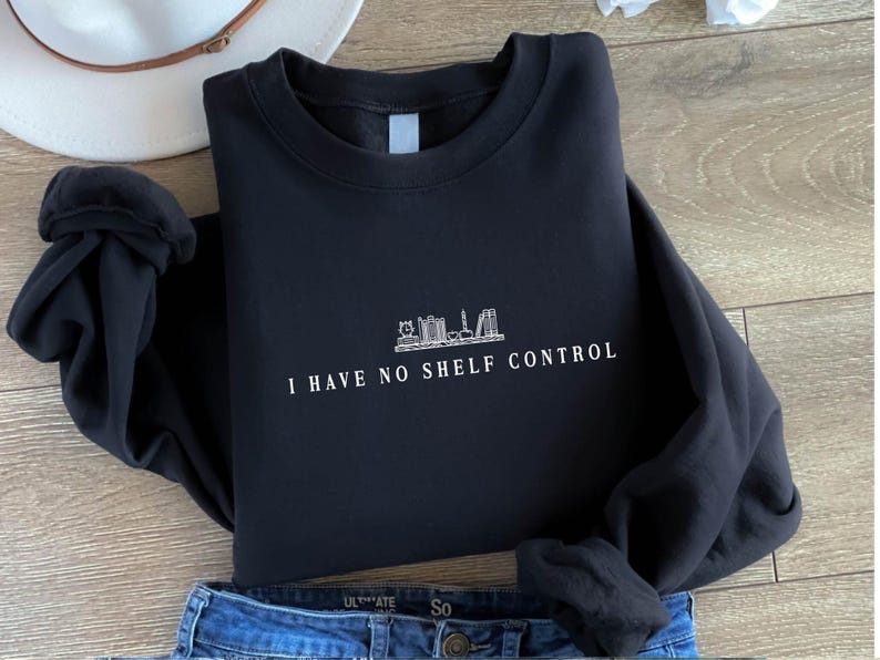 May include: Black sweatshirt featuring the white text "I HAVE NO SHELF CONTROL" and a skyline graphic. The garment is folded, with blue jeans and a white hat in the background. The sweatshirt is made of a soft material.