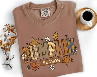 May include: A brown Comfort Colors t-shirt with the words "PUMPKIN SEASON" in a colorful, fall-themed design. The design includes stars, leaves, and floral accents, perfect for autumn.