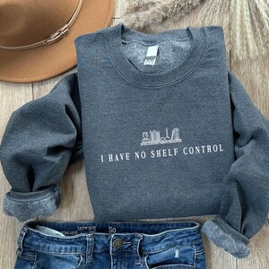 May include: A dark grey sweatshirt with the text "I HAVE NO SHELF CONTROL" in white. The sweatshirt has a crew neck and cuffed sleeves. A brown hat and blue jeans are also in the image.