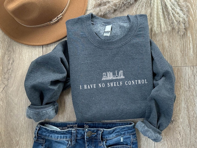 May include: A dark grey sweatshirt with the text "I HAVE NO SHELF CONTROL" in white. The sweatshirt has a crew neck and cuffed sleeves. A brown hat and blue jeans are also in the image.