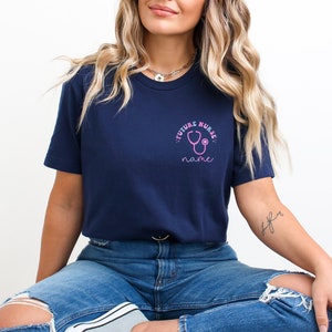 May include: Navy blue t-shirt with a pink stethoscope graphic and the words "FUTURE NURSE" and "name". The shirt is made of a soft material and is suitable for everyday wear. The design is on the upper left chest.
