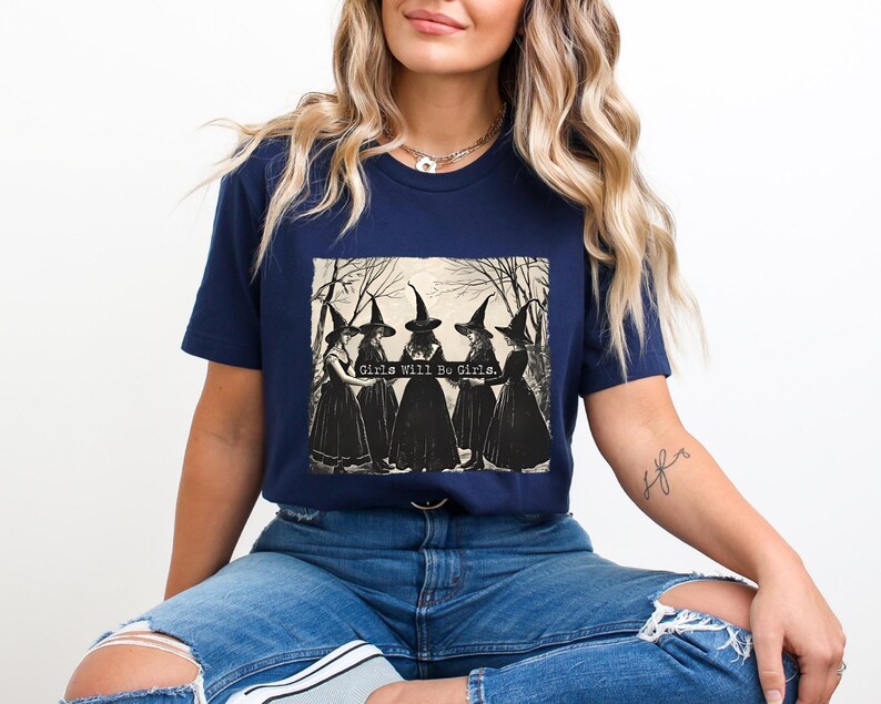 K&ouml;nnte beinhalten: Ein marineblaues T-Shirt mit einem Aufdruck von vier Hexen in schwarzen Kleidern und spitzen H&uuml;ten. Der Text "Girls Will Be Girls" ist auf dem Aufdruck gedruckt.