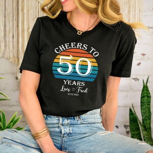 May include: Black t-shirt with a retro sunset graphic and the text "Cheers to 50 Years Luis & Fred 12.02.1950".