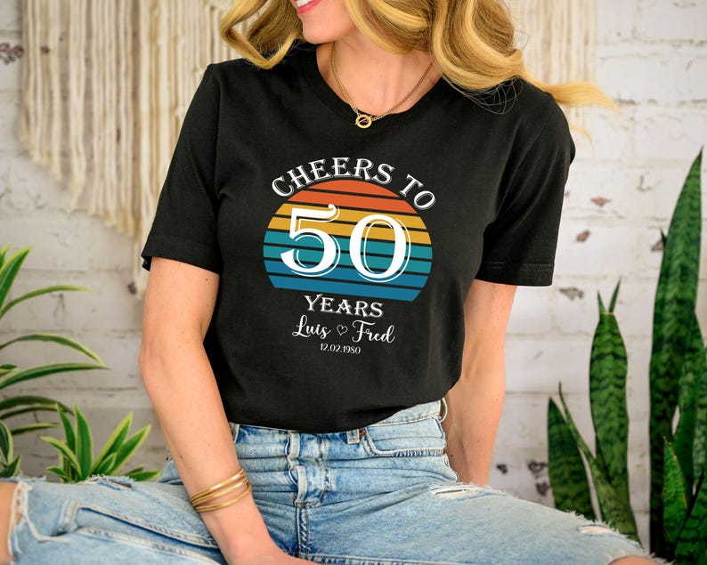May include: Black t-shirt with a retro sunset graphic and the text "Cheers to 50 Years Luis & Fred 12.02.1950".