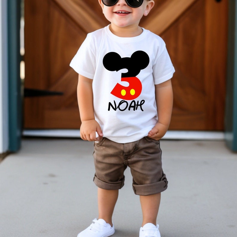 Op de afbeelding: Een wit T-shirt met een Mickey Mouse-ontwerp voor een driejarige. Het ontwerp bevat een zwarte Mickey Mouse-kop, het nummer 3 in rood en de naam "NOAH" in zwart. Het kind draagt een bruine korte broek en witte sneakers.