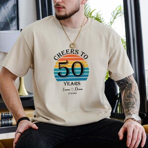 May include: A beige t-shirt with a retro sunset graphic and the text "Cheers to 50 Years Emma &hearts; Dean 12.02.1950".