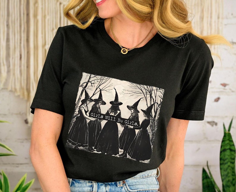K&ouml;nnte beinhalten: Schwarzes T-Shirt mit einem Motiv von vier Hexen in schwarzen Kleidern und spitzen H&uuml;ten, die ein Banner mit der Aufschrift "Girls Will Be Girls." halten.