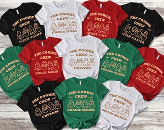 May include: Assorted t-shirts in black, white, red, and green, each featuring the text "The Cookie Crew" with various holiday-themed graphics and phrases like "Flour Power" and "Cookie Maker."