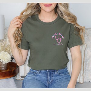 May include: Olive green t-shirt with a pink stethoscope graphic and the words "FUTURE NURSE" and "name". The shirt is a crew neck style and appears to be made of a soft material.