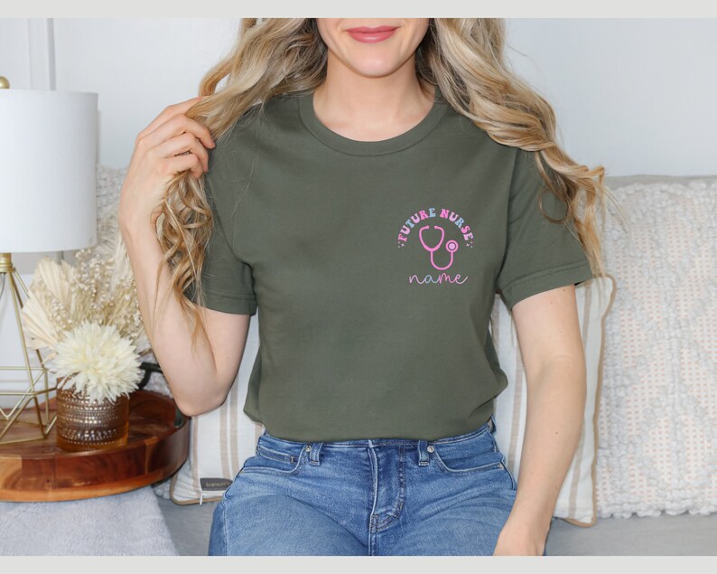May include: Olive green t-shirt with a pink stethoscope graphic and the words "FUTURE NURSE" and "name". The shirt is a crew neck style and appears to be made of a soft material.